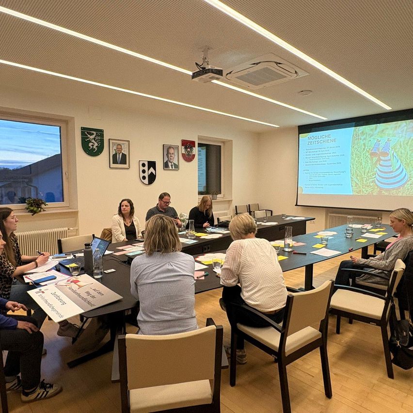 A group of people are in a meeting in a room with a projector screen displaying content, while others take notes on papers.