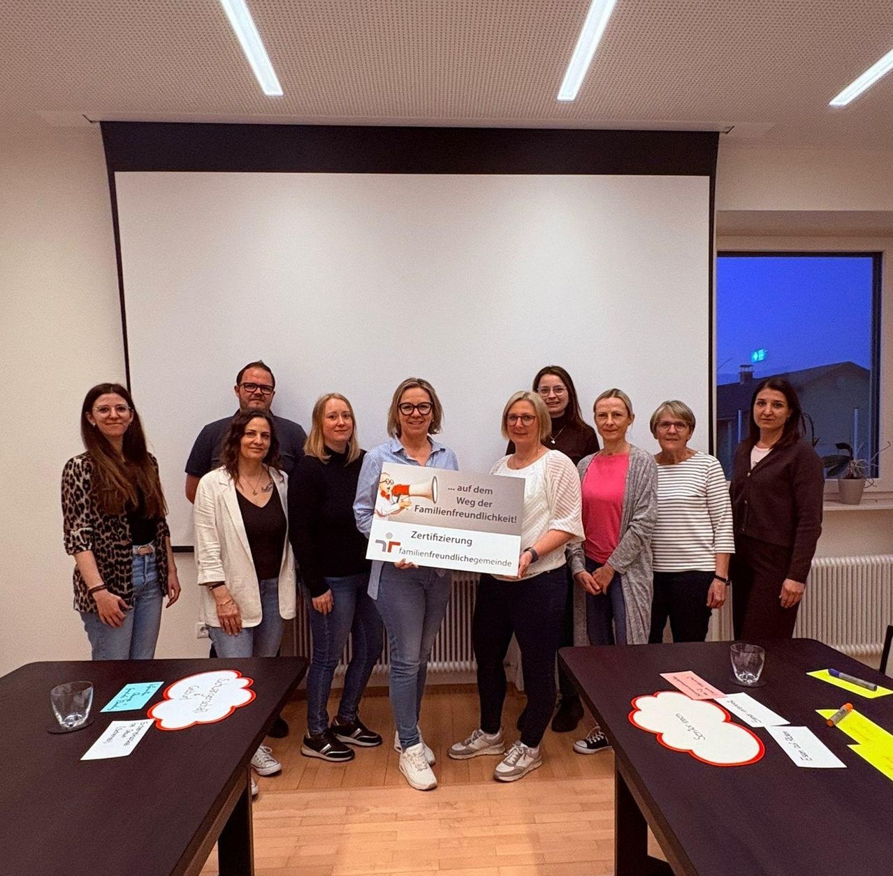 Group of people standing in a conference room with a projector screen. They hold a certificate and a megaphone. The room has two tables with writing on them.