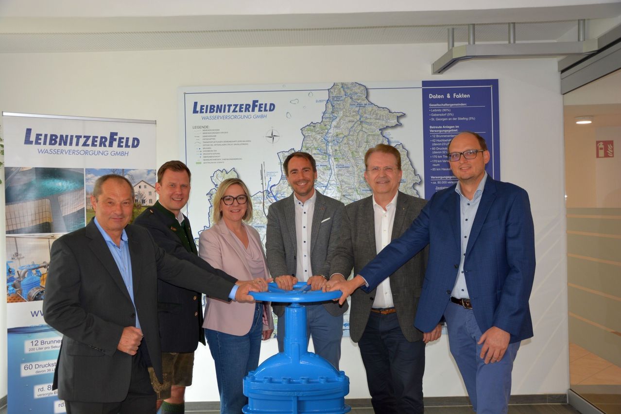 Fünf Personen stehen lächelnd neben einer großen blauen Ventil. Sie stehen vor einer Karte und einem Poster, das Leibnizerfeld Wasserversorgung GmbH sagt.