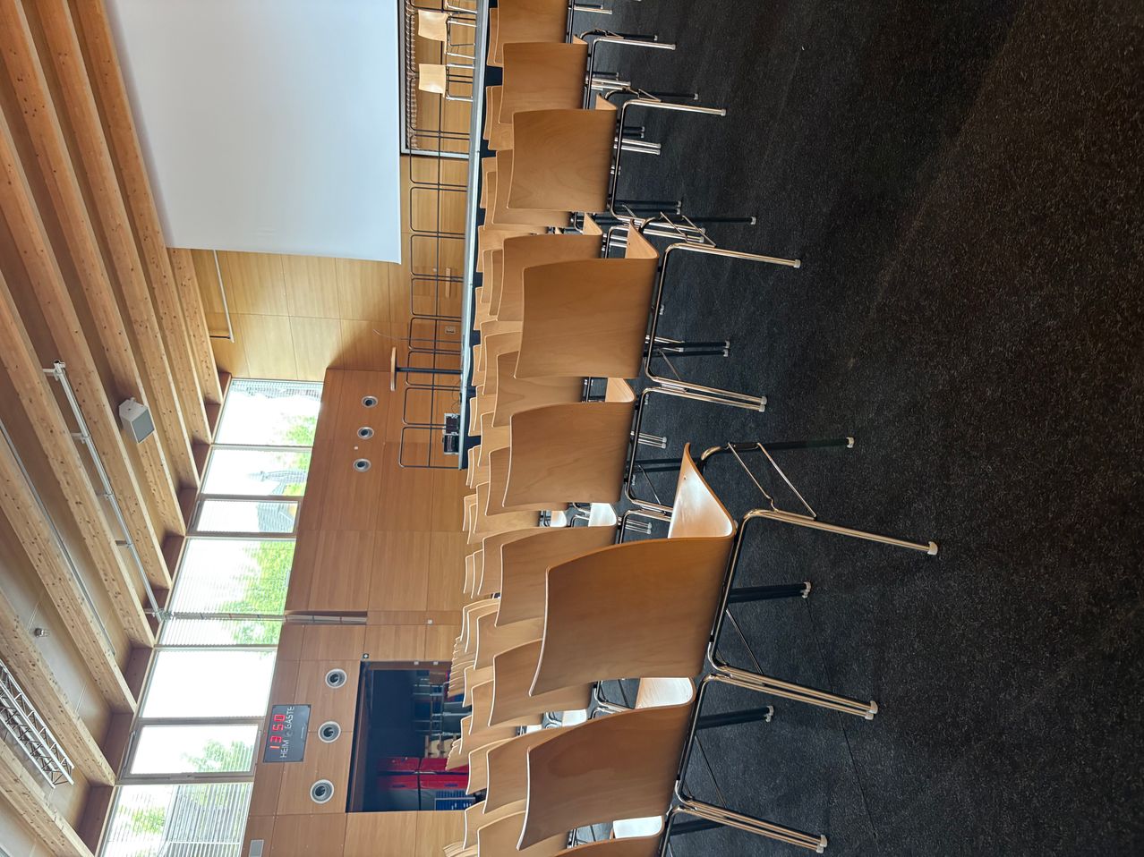 Ein Klassenzimmer mit hölzernen Stühlen und Metallständern, einer weißen Leinwand und Glasfenstern mit einer digitalen Temperaturanzeige.