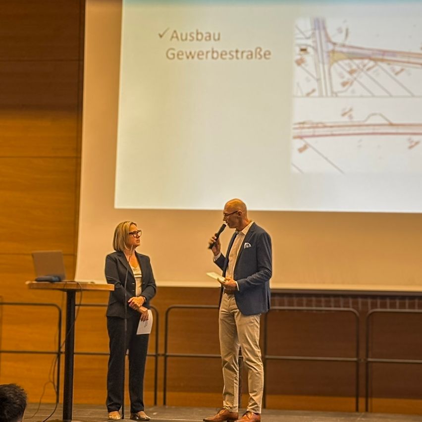 Ein Mann und eine Frau stehen auf einer Bühne. Der Mann hält ein Mikrofon und liest von einem Zettel. Ein Laptop steht auf einem Tisch vor ihnen. Eine Projektionsleinwand zeigt eine Karte mit dem Text 'Ausbau Gewerbestraße'.