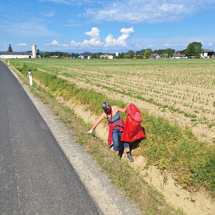 An adult woman collects garbage on the side of the road in a rural area with a wide field behind her.