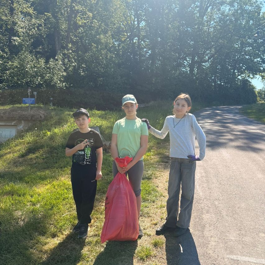 Three kids stand by the roadside, holding a red trash bag. A sunny day with trees in the background.