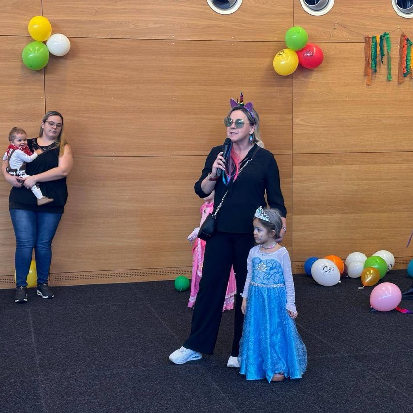 A woman with a unicorn hat speaks into a microphone as a girl in a princess costume stands beside her. A woman holding a baby stands nearby. Balloons are attached to the wall.