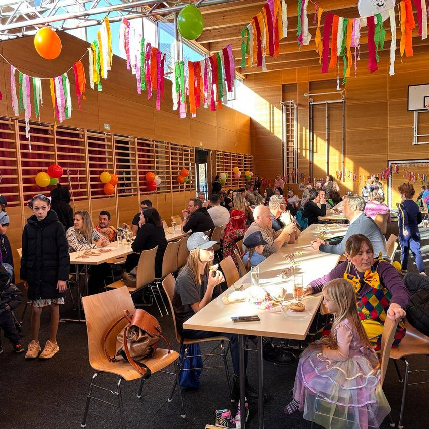 A large group of people are gathered indoors for a party. Some are seated at tables with food, others are standing. Colorful decorations and balloons adorn the room. A girl in a costume stands near a table.