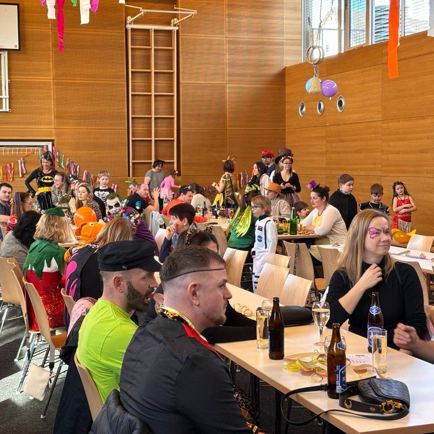 A large group of people in costumes are gathered in a room, seated at tables with drinks and food, and decorations hanging on the wall.