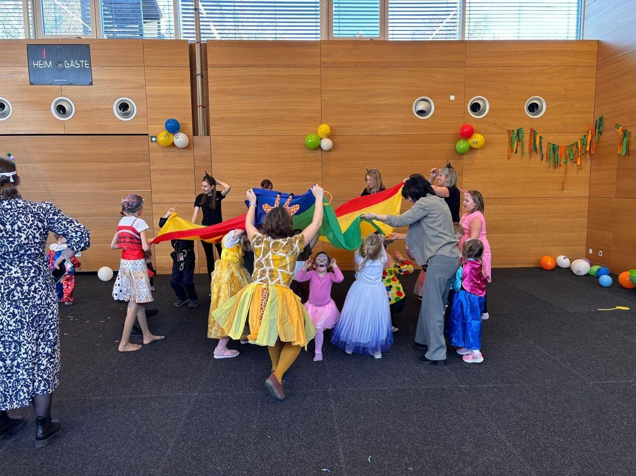 A group of children in costumes are playing with a colorful fabric in a room with a wooden wall. Some adults are watching and participating.
