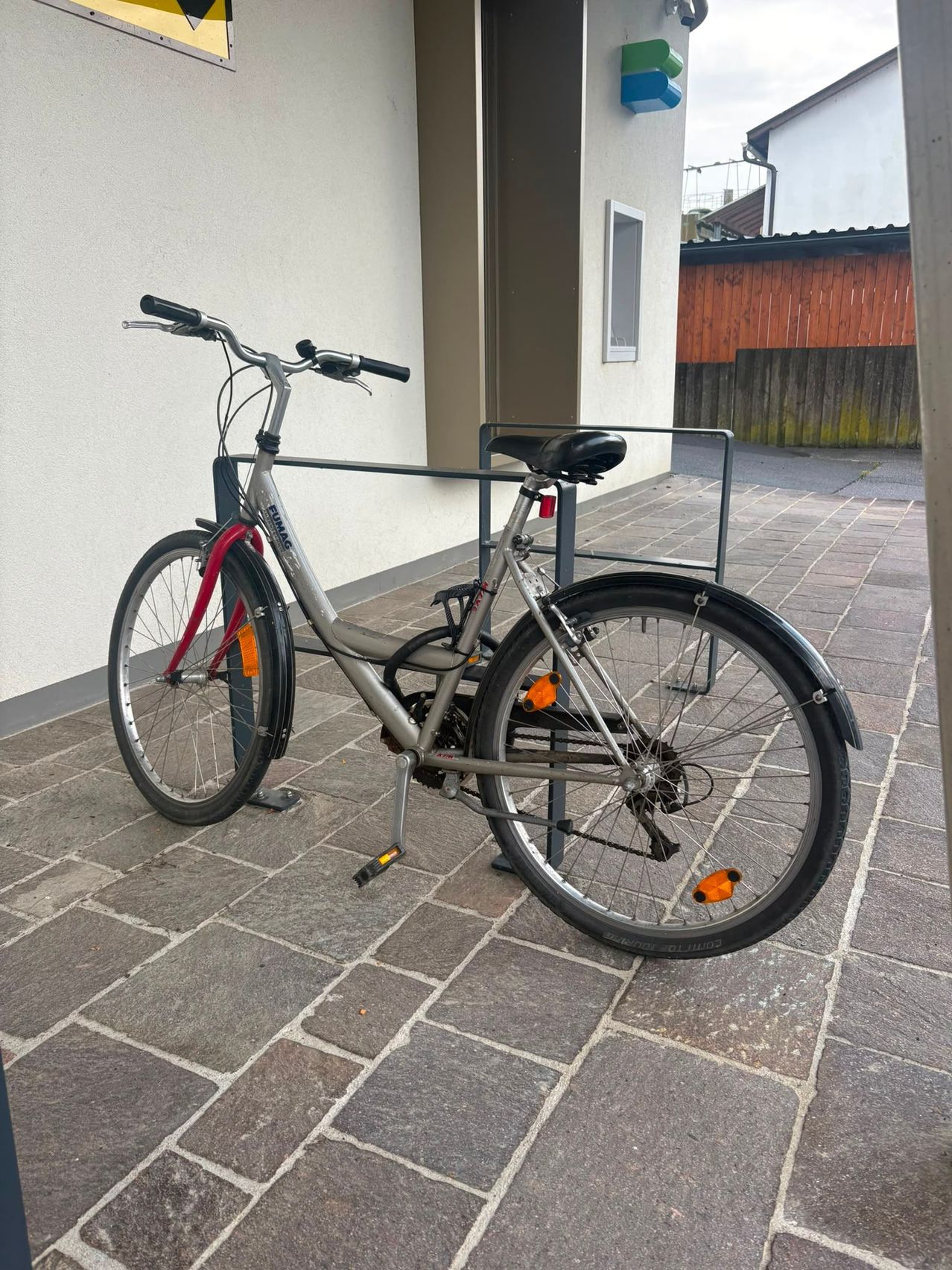 Ein silbernes Fahrrad mit roten Akzenten ist auf einem gepflasterten Bürgersteig neben einem Gebäude geparkt. Das Fahrrad hat orangefarbene Reflektoren an den Rädern.
