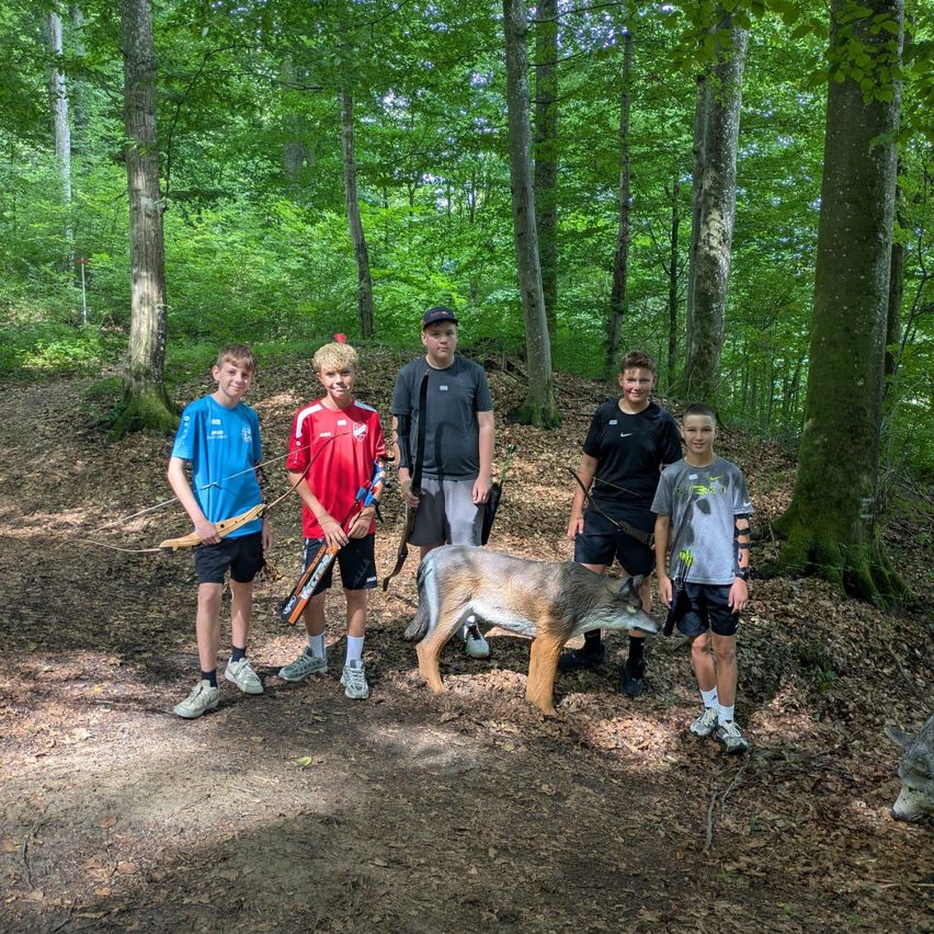Fünf Jungen stehen mit Bögen und Pfeilen in einem Wald und posieren für ein Foto. Vor ihnen steht eine Wolfsstatue.