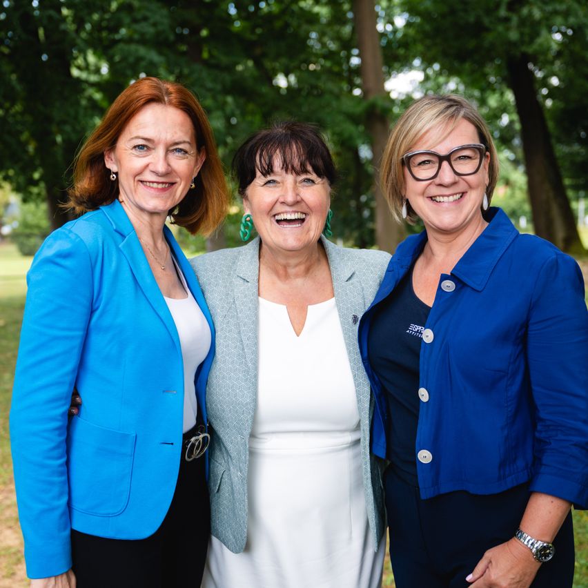 Drei Frauen stehen lächelnd in einem Park und sind in professioneller Kleidung gekleidet. Die Frau links trägt einen blauen Blazer, die mittlere Frau einen grauen Blazer und die Frau rechts einen blauen Blazer mit einem Logo auf der linken Seite.