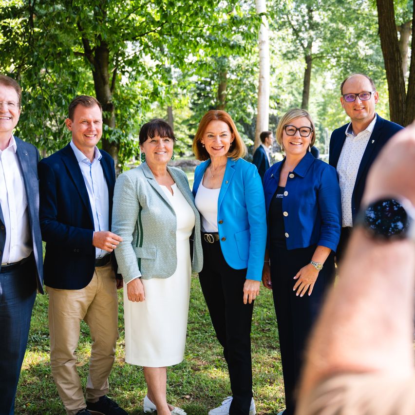Eine Gruppe von sechs Personen, in formeller Kleidung gekleidet, steht zusammen in einem Park und lächelt für ein Foto. Die Frau in der Mitte trägt ein weißes Kleid und eine hellgrüne Jacke.