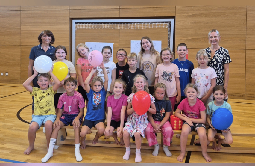 Eine Gruppe von Kindern und zwei Erwachsenen posiert für ein Foto in einer Turnhalle mit Luftballons. Die Kinder sitzen auf einer Bank, und die Erwachsenen stehen hinter ihnen. Die Kinder halten Luftballons, und einige tragen Brillen. Die Turnhalle hat Holzwände und ein Netz im Hintergrund.