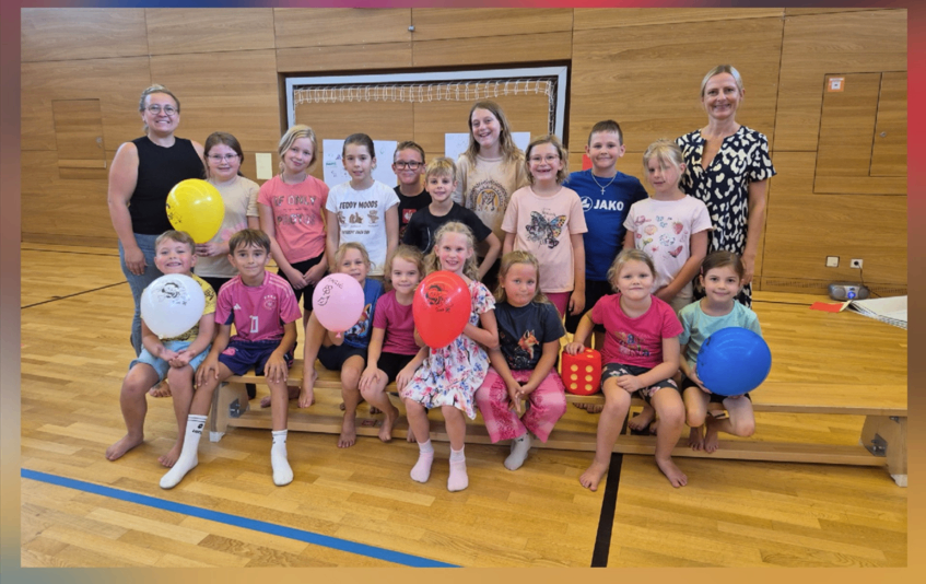Eine Gruppe von Kindern und Erwachsenen posiert für ein Foto in einer Turnhalle. Die Kinder halten Luftballons und sitzen auf einer Bank. Die Erwachsenen stehen hinter ihnen.