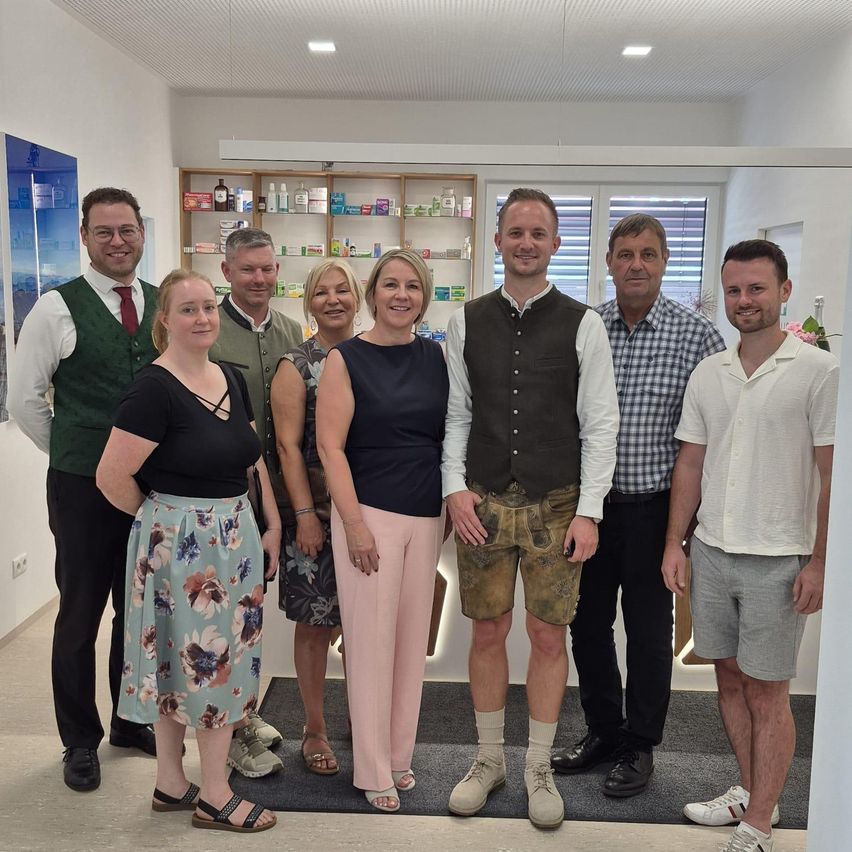 A group of people stands in a room, smiling for a photo. Two women in dresses, three men in various attire, and a woman in a green vest. Shelves with items and a window with blinds.