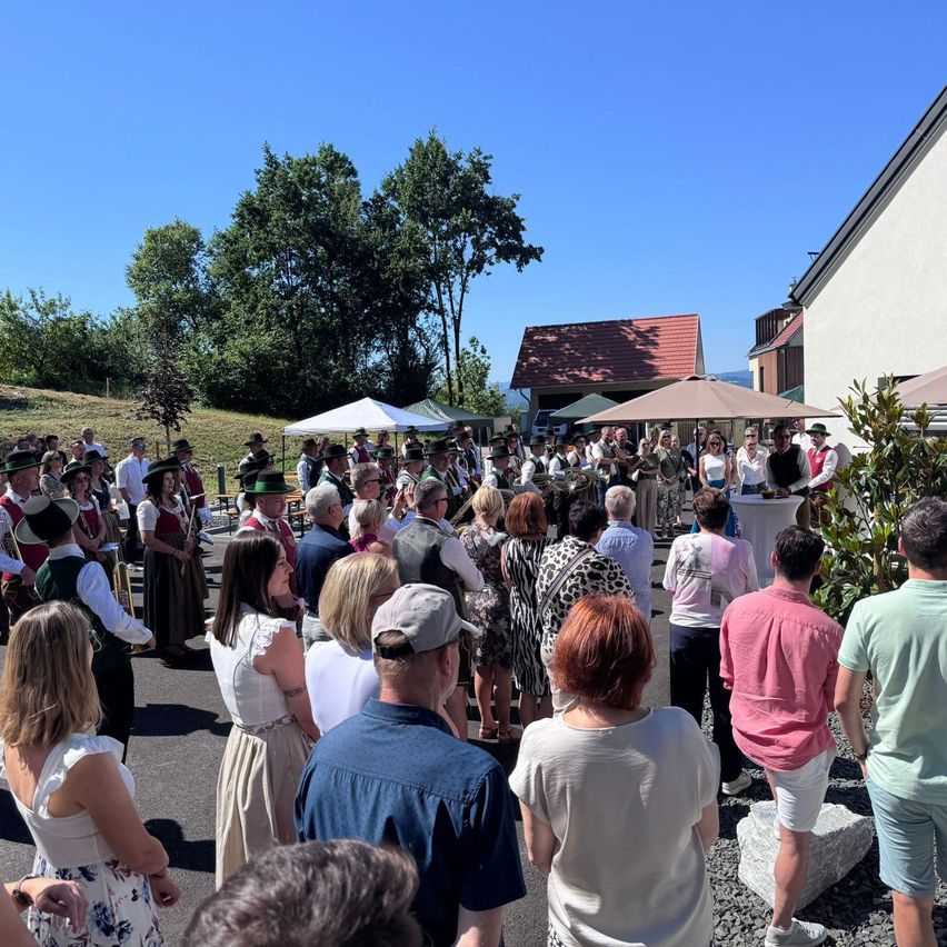 A crowd of people gathers outside a building with a red roof, some wearing hats. Nearby, there are umbrellas and tents, and the setting is sunny.