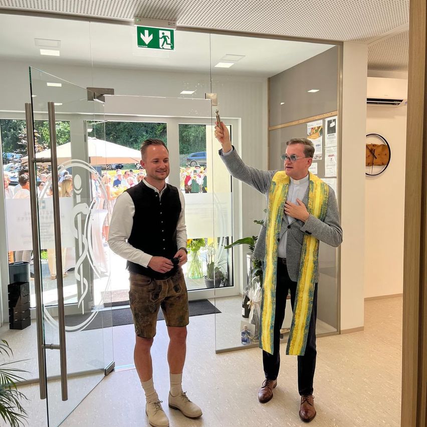 Two men stand in front of a glass door. One holds a key, the other wears a vest. They smile at the camera. An emergency exit sign is above the door.