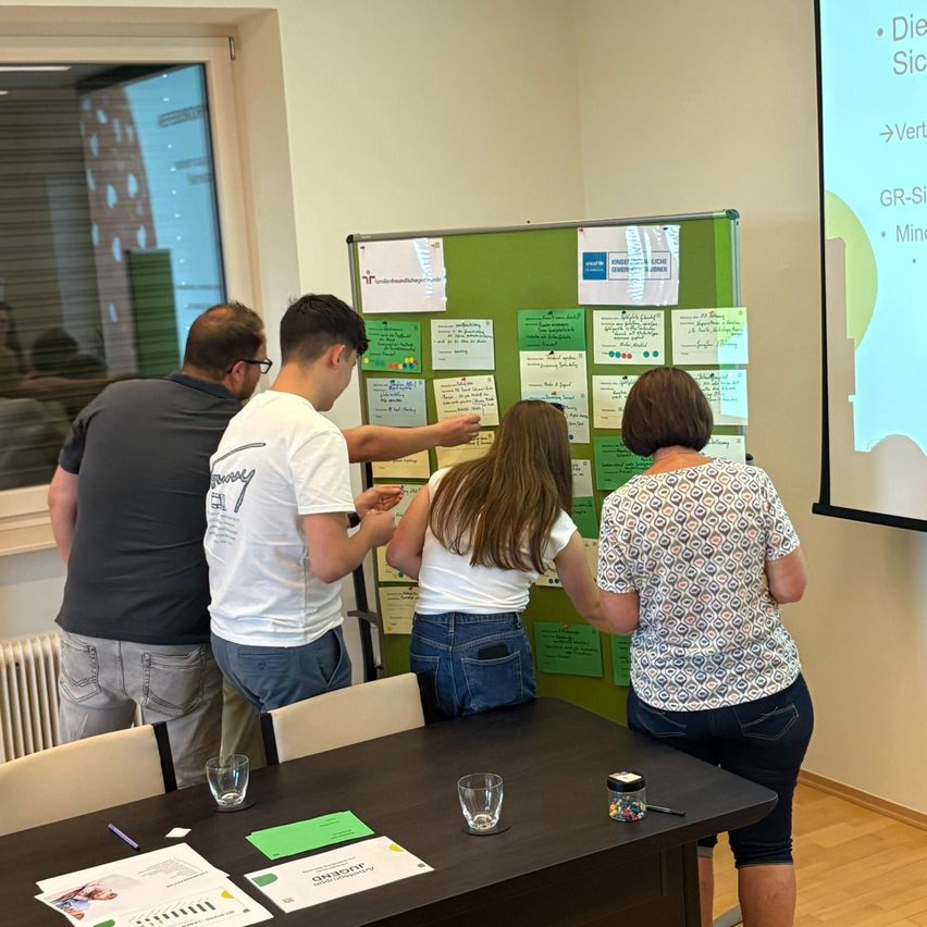 A group of people stand around a green board with sticky notes. Two women and two men are looking at the board. One man points at a sticky note.