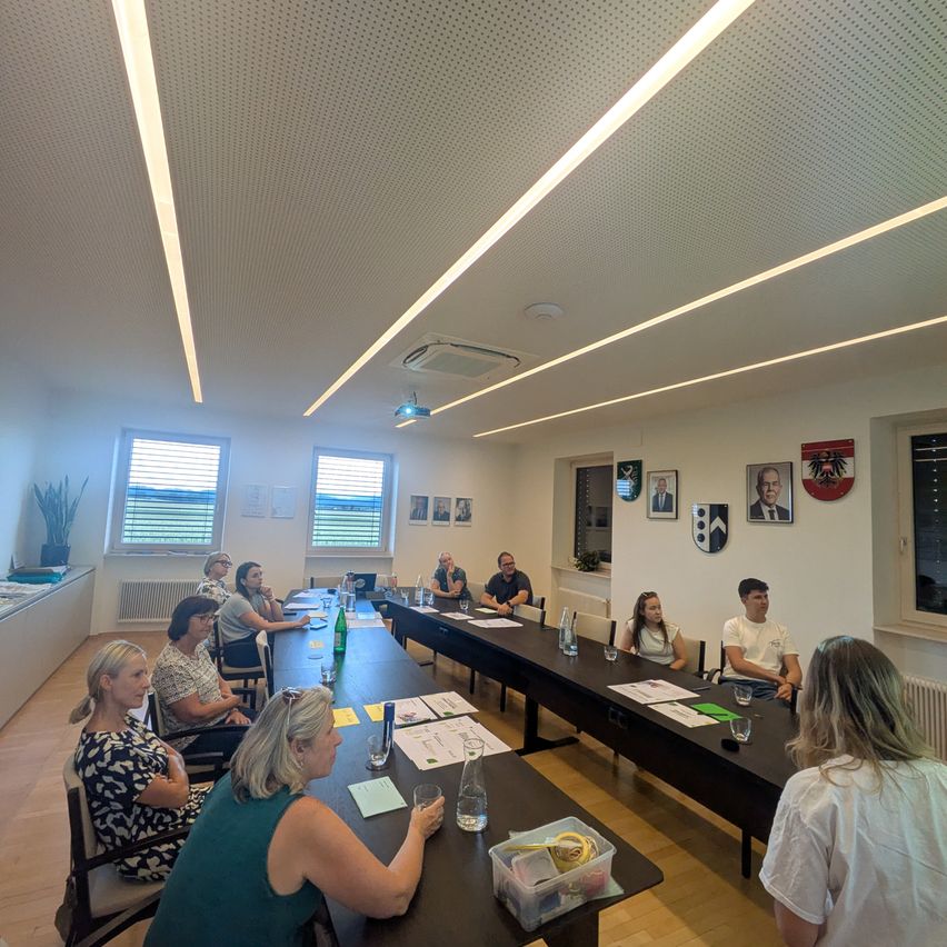 A group of people are sitting around a long table in a meeting room. The room is well-lit, with white walls and a ceiling. Some of them are engaged in conversation, while others are listening attentively.