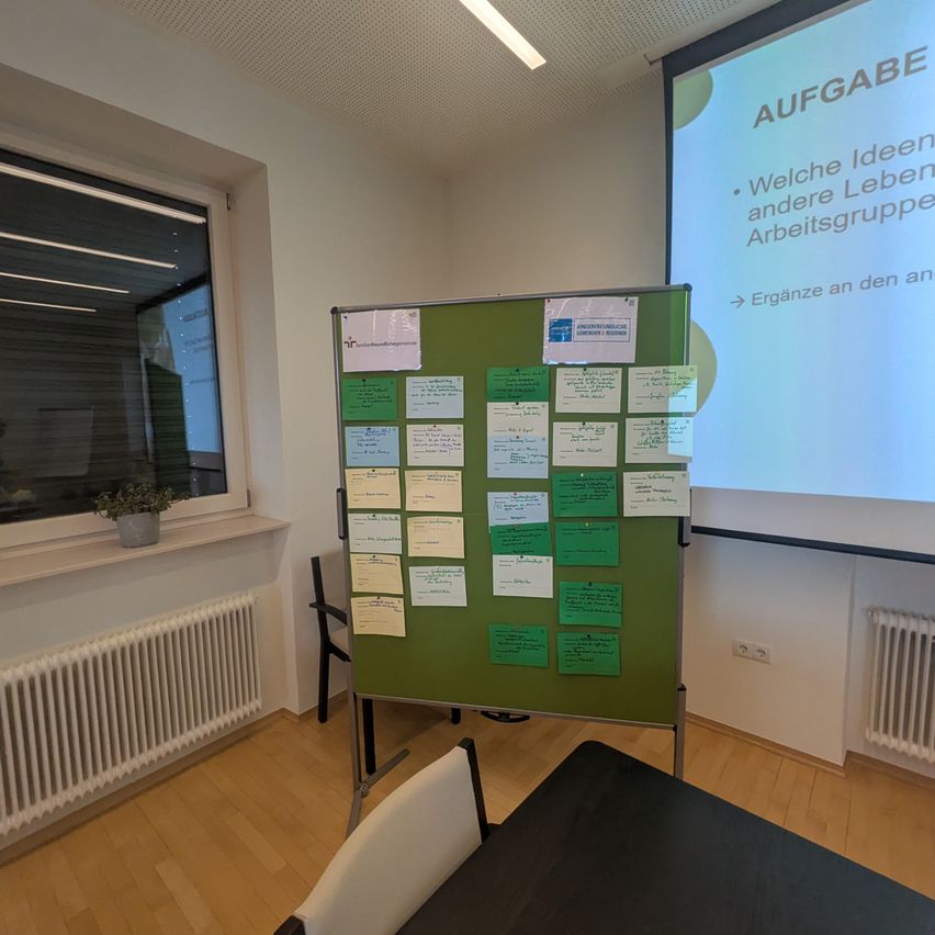 An office with a bulletin board covered in green sticky notes. A projector screen in the background displays a slide with blue text.