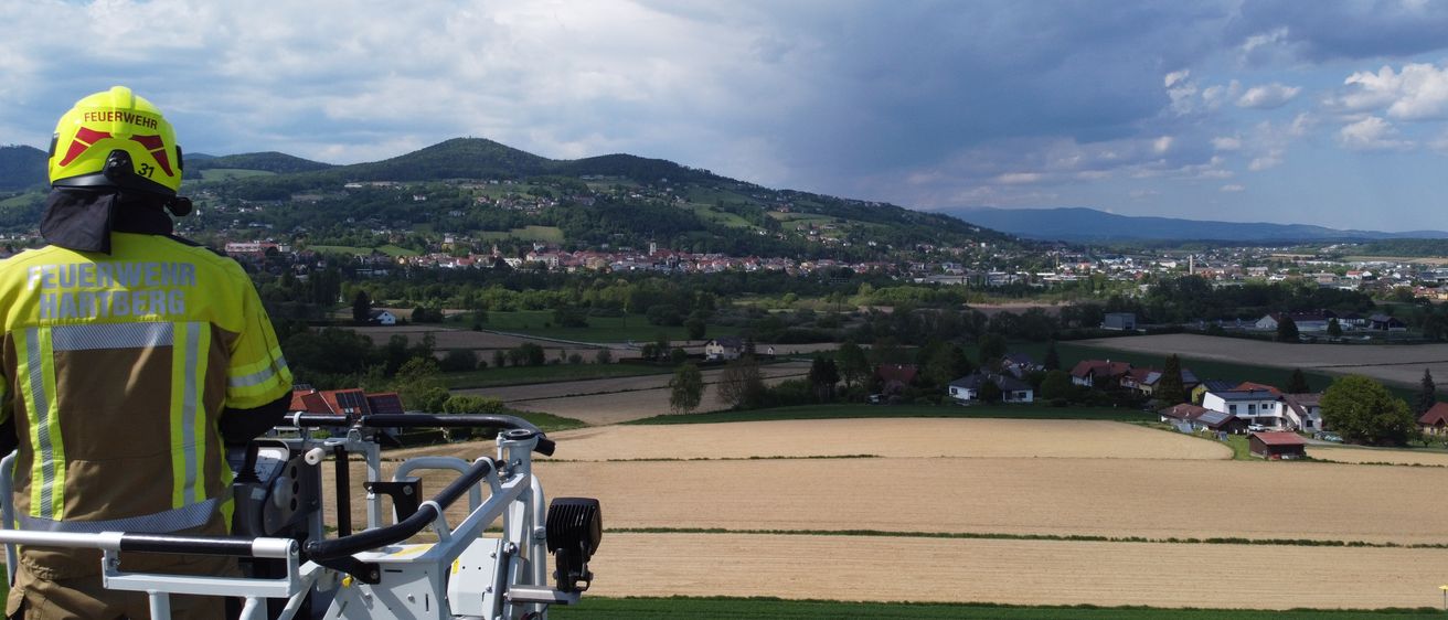 Ein Feuerwehrmann steht in einem Kran und überblickt eine ländliche Landschaft mit grünen Feldern, entfernten Bergen und einer Stadt.
