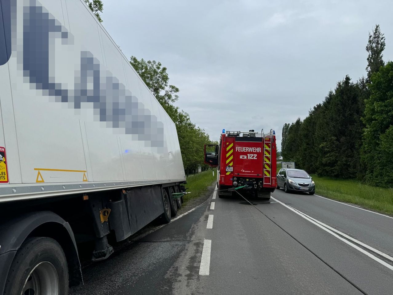 Ein roter Feuerwehrwagen steht am Straßenrand neben einem Lkw und einem silbernen Auto dahinter. Der Lkw hat ein großes Logo an seiner Seite.