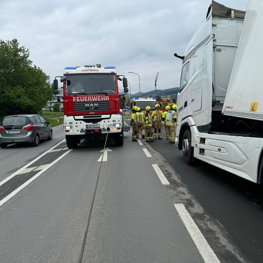 Ein roter Feuerwehrwagen und ein weißer Lkw stehen auf der Straße. Eine Gruppe von Feuerwehrleuten steht an der Seite, trägt gelbe Helme und Stiefel. Ein silberner Wagen fährt auf der Straße. Bäume und Gebäude sind in der Ferne.