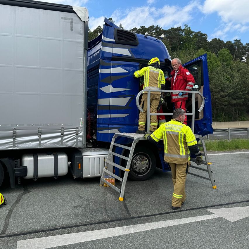Zwei Feuerwehrleute in gelben Uniformen steigen auf eine Leiter zum Führerhaus eines blauen Lastwagens. Ein Mann in einem roten Jackett steht neben der offenen Tür.
