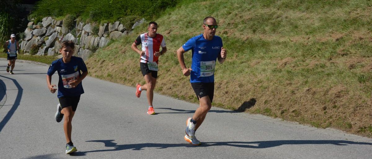 Drei Läufer joggen auf einer Straße mit einem grasbewachsenen Hügel an der Seite. Der Mann in Blau führt, gefolgt von zwei anderen. Der Boden ist teilweise mit Gras und Erde bedeckt.