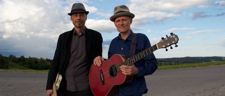 Zwei Männer, beide mit Hüten, stehen mit Gitarren lächelnd auf einer Straße unter einem bewölkten Himmel.