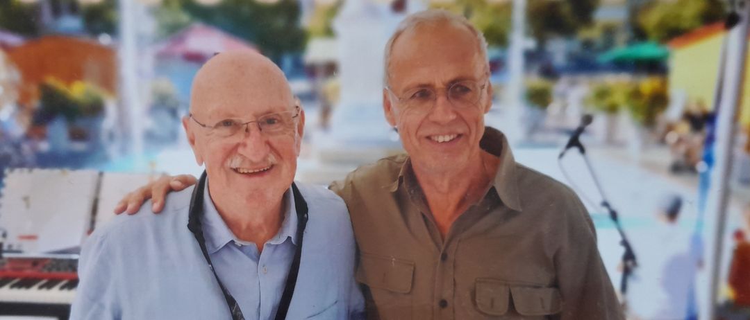 Zwei ältere Männer stehen lächelnd vor einer Kulisse mit Pool und Musikinstrumenten.