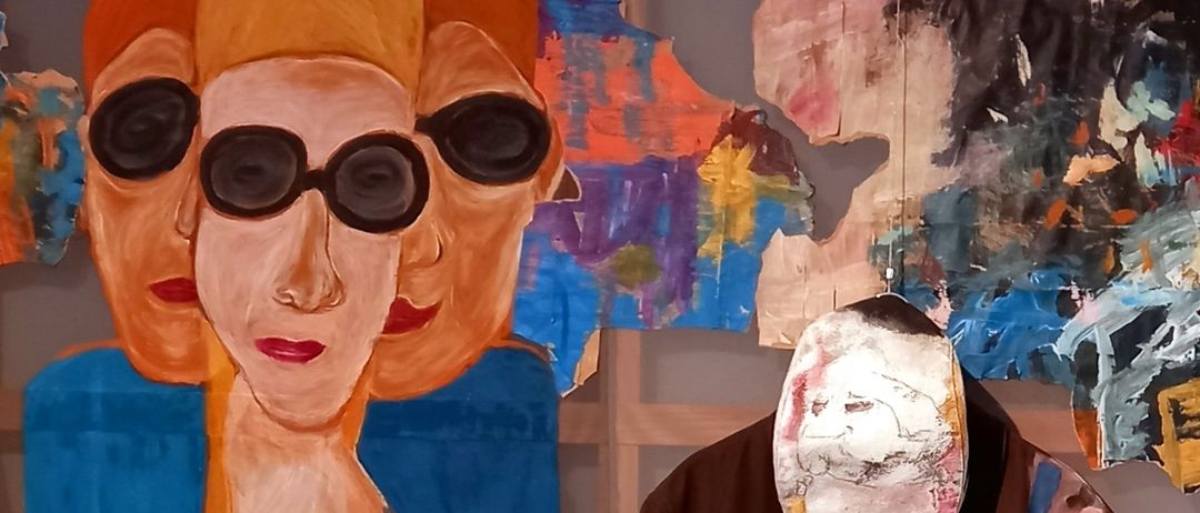 Ein abstraktes Gemälde einer Frau mit rotem Lippenstift und Sonnenbrille hängt an einer Wand. Daneben ist ein farbenfrohes, abstraktes Kleidungsstück mit Gesichtsmuster ausgestellt.