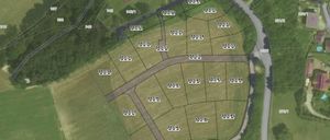 Eine Luftaufnahme eines ländlichen Gebiets mit verschiedenen Grundstücken, die jeweils mit einer Nummer gekennzeichnet sind. Straßen und Wege führen zu einem zentralen Bereich. Grüne Felder und Bäume umgeben die Grundstücke.