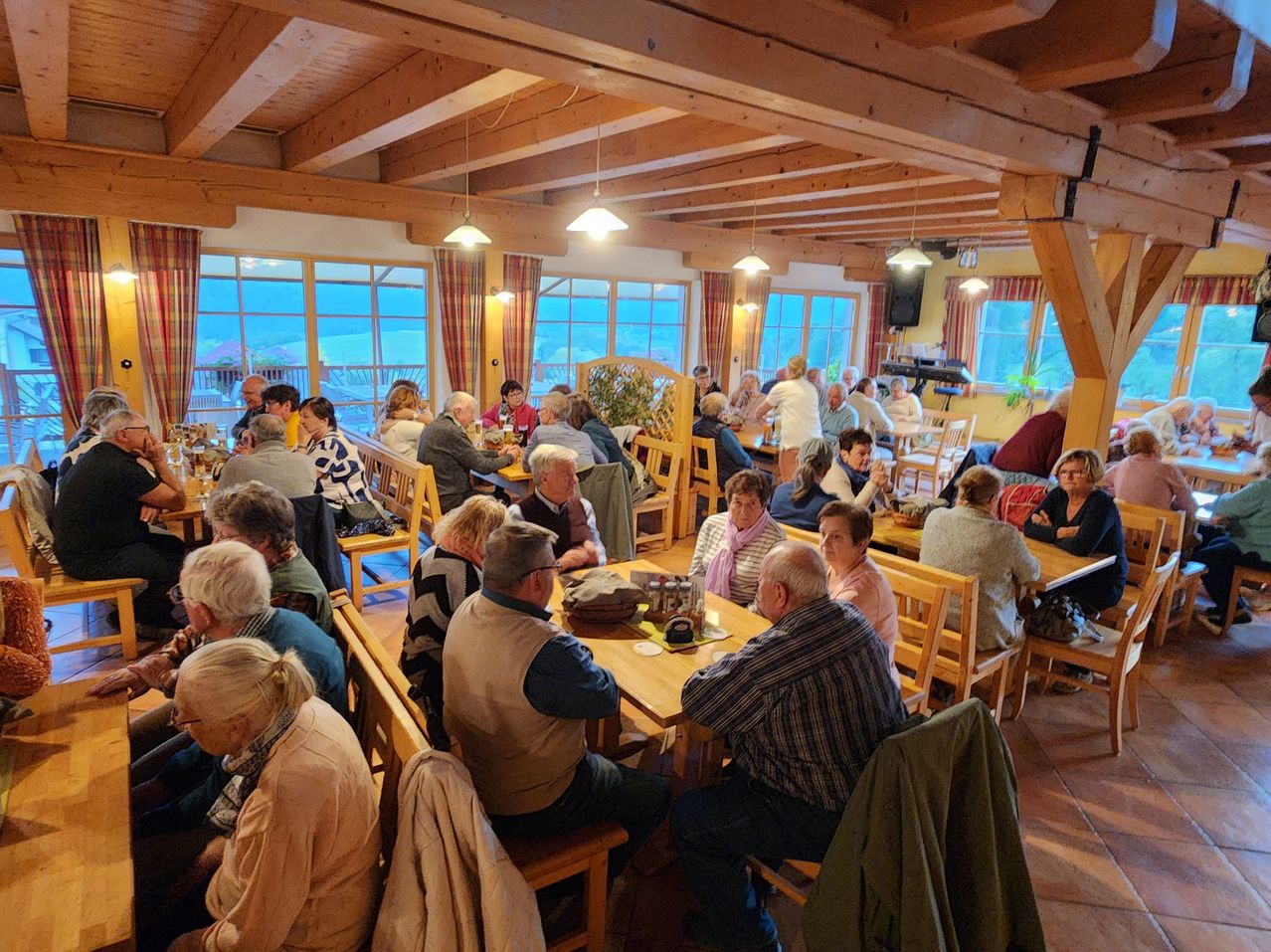 A dining room with wooden ceiling beams and large windows. People are seated at tables, some eating, others conversing. The room is filled with natural light.