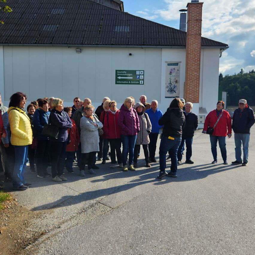 A group of people stands outside a building with a green sign and brick chimney. The building has white walls and a brown roof.
