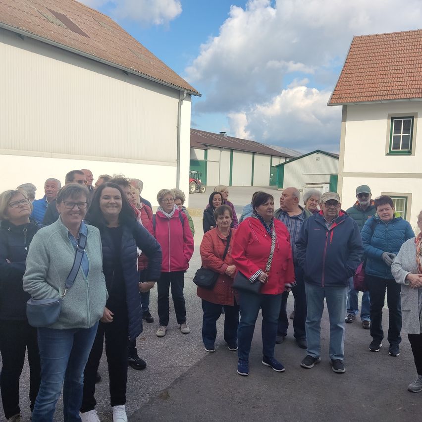 A group of people in winter clothing are standing outside a building with a tractor in the background. The weather is cloudy.