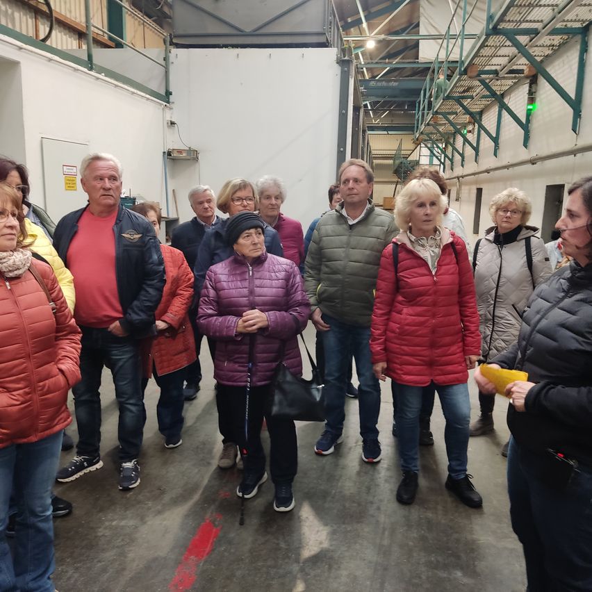 A group of people stand in a corridor of a warehouse. They are all dressed warmly in winter clothing. A woman holding a cane leads the group.