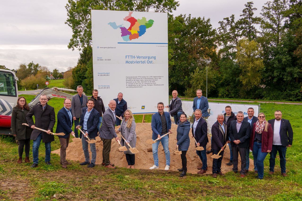 A group of people, dressed in formal attire, stand in front of a large sign and hold shovels. They are participating in an event with a sign reading FTTH-Versorgung Moestviertel Ost. Behind them, a grassy area and trees are visible.