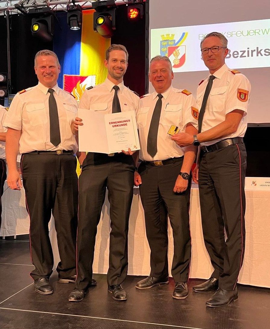 Vier Männer in formellen Uniformen stehen auf einer Bühne. Die zentrale Figur hält ein Zertifikat. Sie scheinen eine Auszeichnung zu feiern. Hinter ihnen ist ein mit einem weißen Tuch bedeckter Tisch und eine Leinwand mit einem Logo zu sehen.