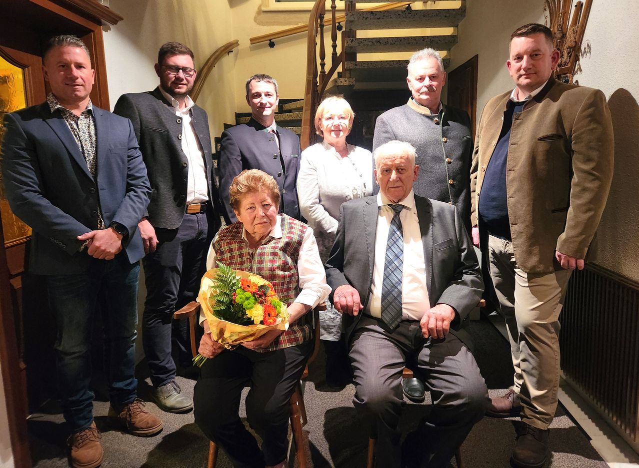 Ein älteres Paar sitzt auf einem Stuhl und hält einen Blumenstrauß, umgeben von anderen Menschen in formeller Kleidung, möglicherweise bei einer Feier.