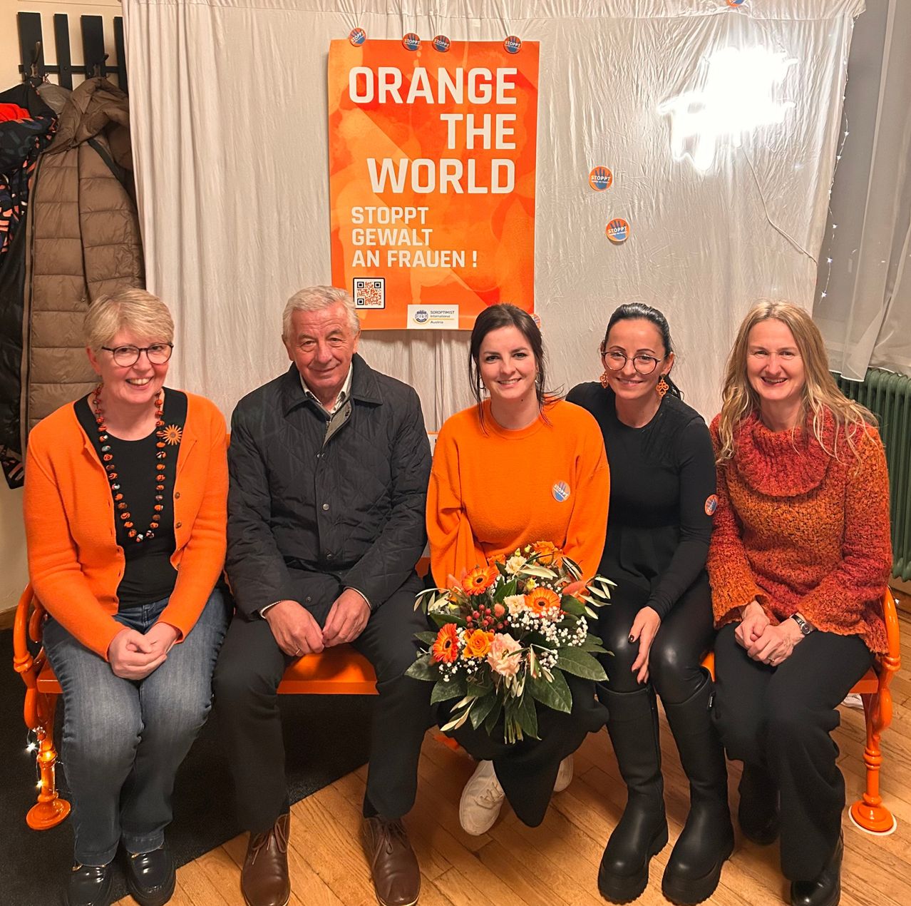 Fünf Personen sitzen auf einer orangefarbenen Bank vor einem weißen Vorhang. Sie tragen orange Kleidung. Eine Frau hält einen Blumenstrauß. Hinter ihnen steht ein Banner mit der Aufschrift 'Orange the World'.