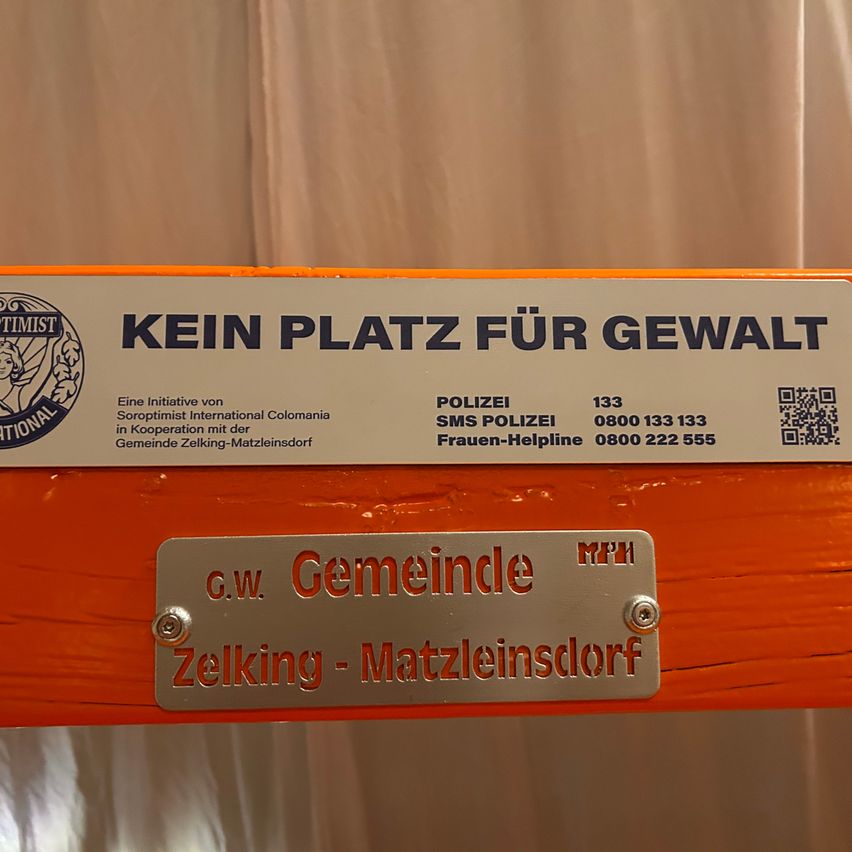 Ein orangefarbenes Schild mit einem Logo trägt die Aufschrift 'KEIN PLATZ FÜR GEWALT'. Darunter sind Kontaktdaten für Polizei und Frauen-Hotline. Ein silbernes Schild darunter sagt 'G.W. Gemeinde' mit dem Stadtnamen 'Zelkíng - Matzleinsdorf'.