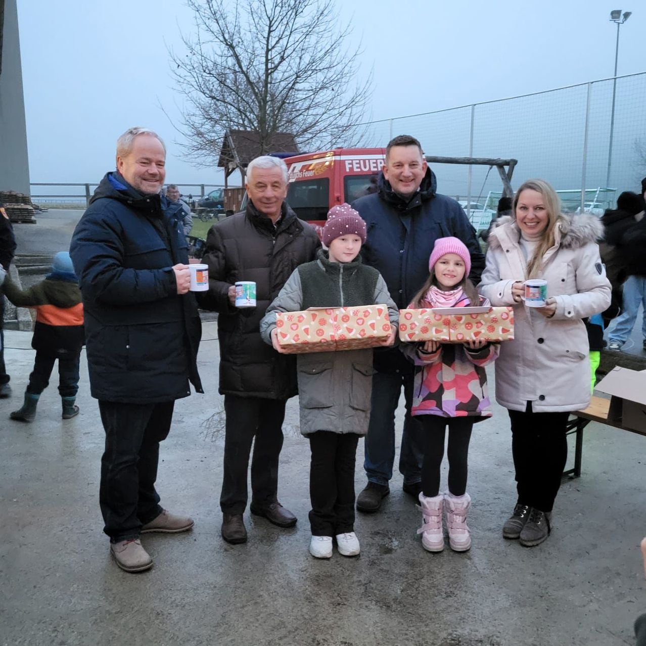 Eine Gruppe von Erwachsenen und Kindern steht zusammen, hält Geschenke und Tassen und lächelt für ein Foto. Dahinter ist ein rotes Fahrzeug geparkt.