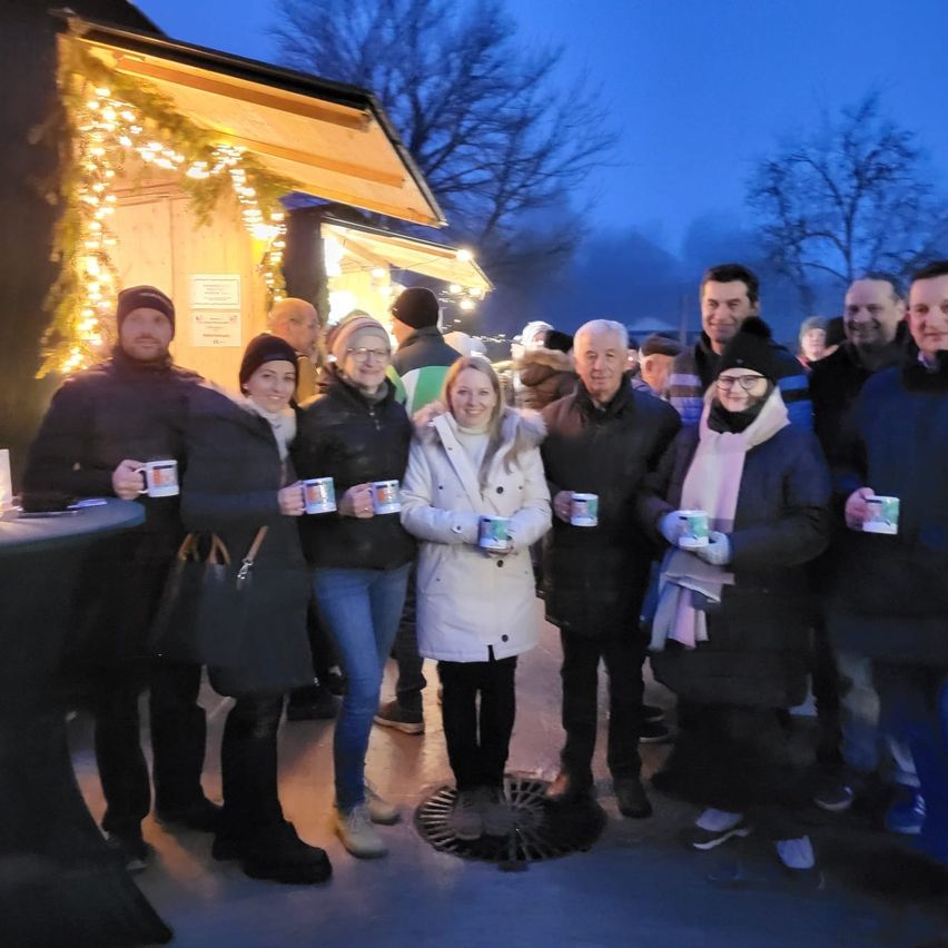 Eine Gruppe von Menschen in Winterkleidung steht draußen, lächelt und hält Tassen in der Hand. Sie stehen vor einem Gebäude, das mit Weihnachtslichtern und Bäumen geschmückt ist.