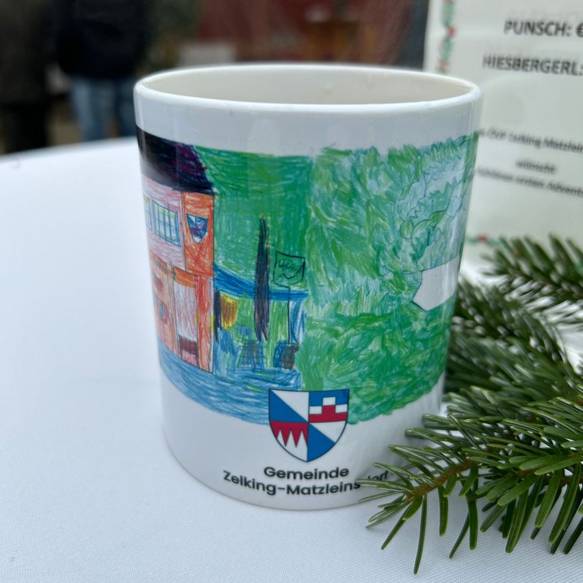 Ein Becher liegt auf einer weißen Tischdecke. Er hat eine farbenfrohe Illustration eines Hauses, Bäume und eines Schildes. Die Worte 'Gemeinde Zelking-Matzleinsdorf' sind am unteren Rand geschrieben. Ein Tannenzweig befindet sich auf der rechten Seite.