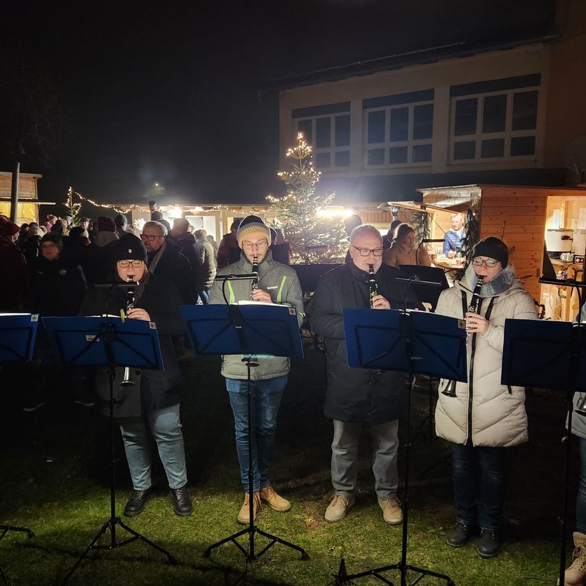 Eine Gruppe von Musikern steht auf einem Rasen und spielt Klarinetten mit Notenständern vor sich. Ein Publikum schaut im Hintergrund zu, und ein Weihnachtsbaum ist mit Lichtern geschmückt.