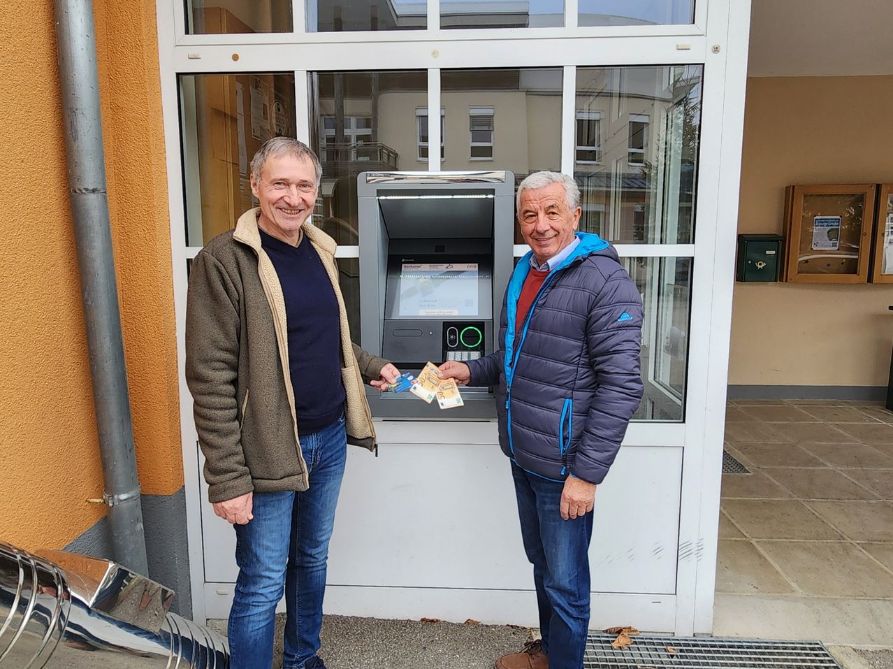 Zwei Männer stehen lächelnd vor einem Geldautomaten. Der Mann auf der linken Seite hält Bargeld, und der Mann auf der rechten Seite hält einen Quittungsbeleg. Sie tragen beide Jacken und Jeans. Hinter ihnen ist eine Glastür mit einem Gebäude im Inneren sichtbar.
