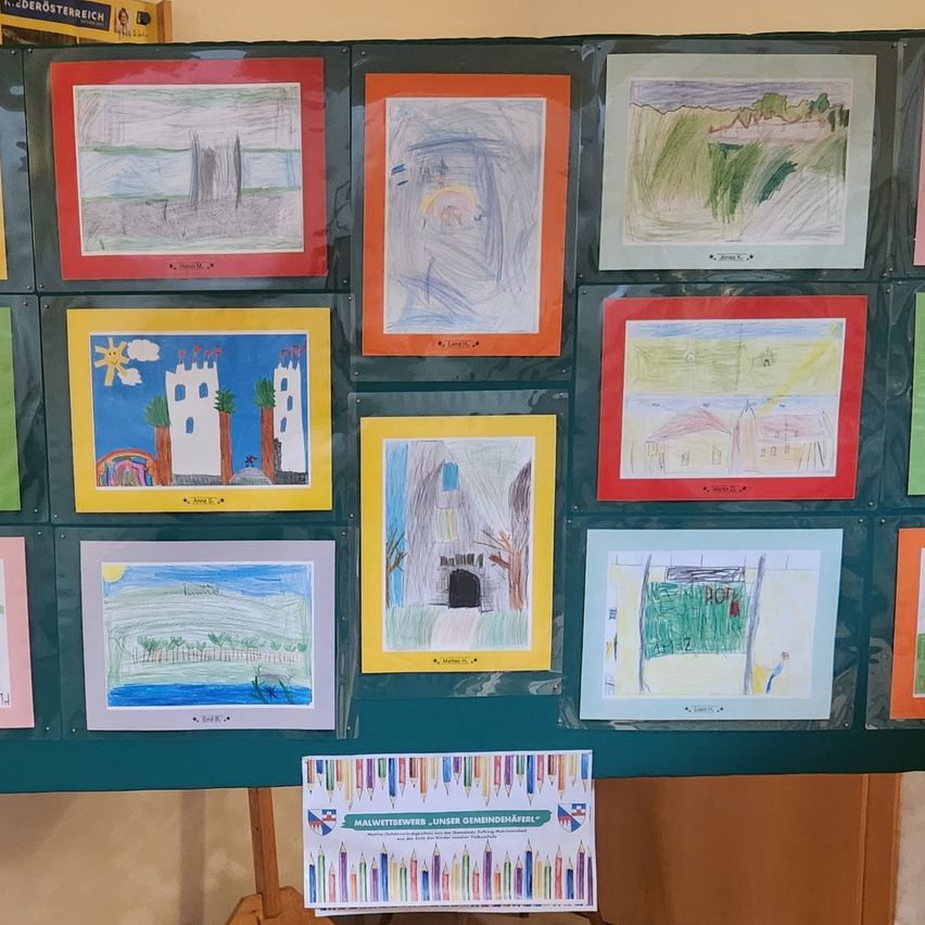 Eine grüne Wand mit mehreren gerahmten Zeichnungen von Kindern. Einige Zeichnungen zeigen Landschaften und Gebäude, andere abstrakte Designs. Ein Schild am unteren Rand zeigt an, dass es Teil eines Malwettbewerbs ist.