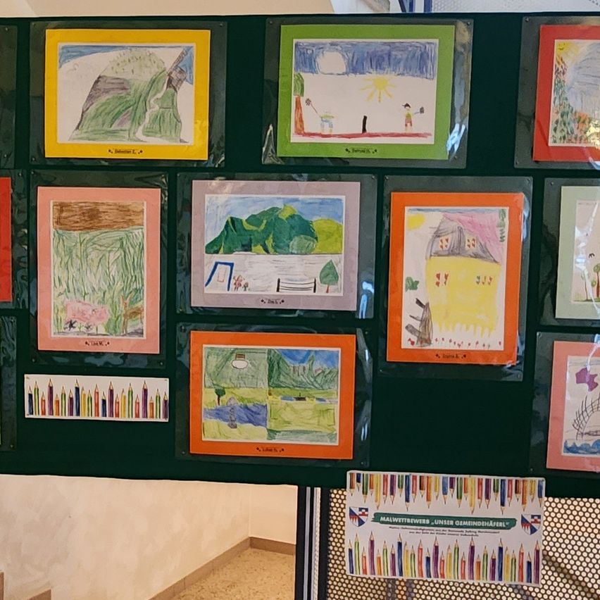 An einer Wand sind mehrere gerahmte Zeichnungen in verschiedenen Farben angebracht. Die Kunstwerke umfassen Landschaften, Häuser und abstrakte Designs. Unten sind eine kleine Reihe bunter Stifte zu sehen. Ein Schild unten links trägt die Aufschrift 'Maiwettbewers_Unser Gemeindehaeferl'.