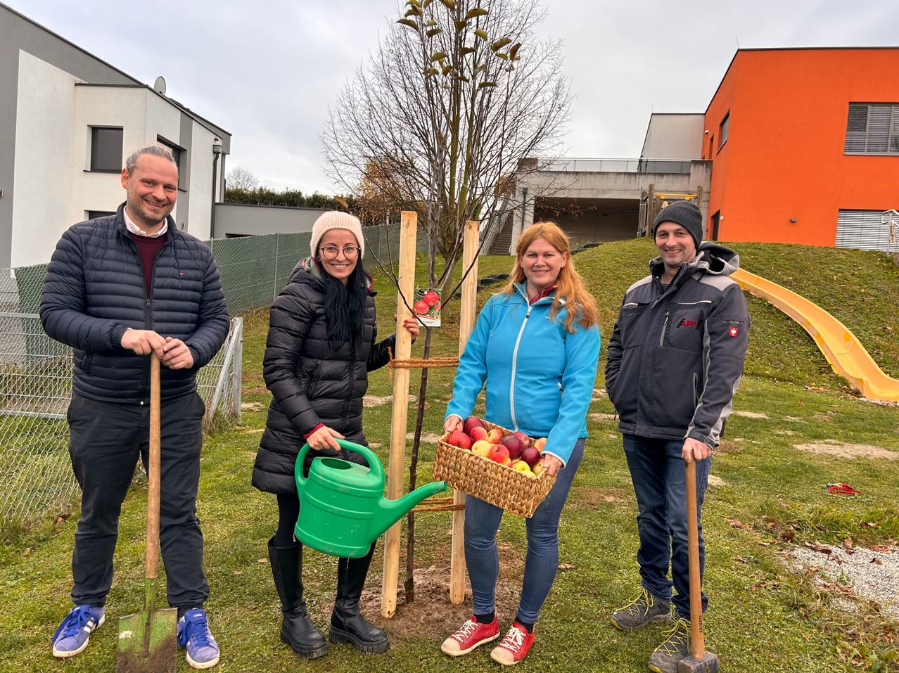 Four people are standing on a grassy field near a tree, two holding shovels and two holding watering cans and a basket of apples. Behind them, there is a building with an orange wall.