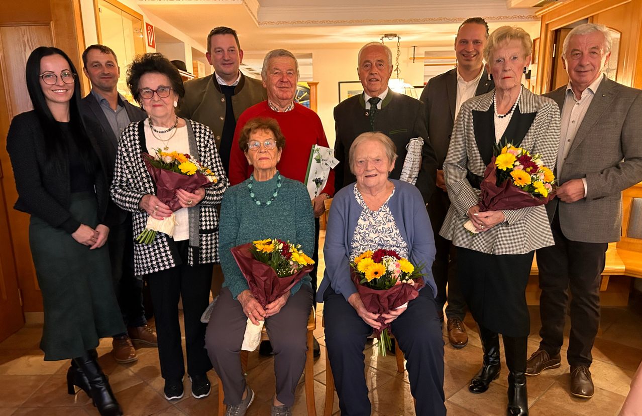 Eine Gruppe älterer Menschen und jüngere Erwachsene posiert drinnen für ein Foto, alle mit Blumensträußen. Die älteren Frauen sitzen auf Stühlen, während andere dahinter stehen.
